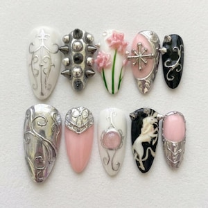 May include: A set of ten press-on nails featuring diverse designs including silver filigree on white, metallic studs, pink floral accents, a silver cross, black with silver swirls, a solid silver finish, pink with silver armor, and a unicorn graphic.