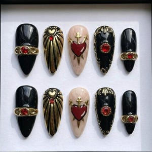 May include: A set of ten press-on nails in a black box. The nails feature various designs, including black, gold, and nude colors. Some nails have gold accents, red gems, and heart and cross motifs. The nails are oval and almond-shaped.