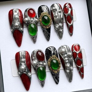 Può includere: Un set di dieci unghie finte lunghe e appuntite. Le unghie presentano una varietà di design, tra cui glitter rossi, accenti metallici argentati e abbellimenti di gioielli in rosso, verde e oro. Le unghie sono esposte in una scatola nera.