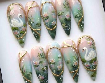 May include: A set of ten long, almond-shaped artificial nails. Each nail features a hand-painted design of a swan, water lilies, and a pond scene, framed with gold accents and small white pearls. The color palette includes pink, green, and white.