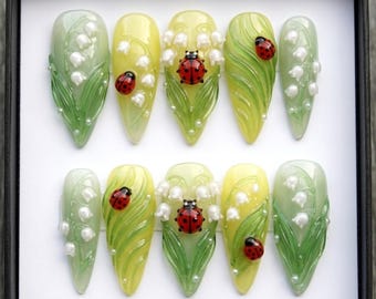 May include: A set of ten artificial nails, each with a unique design. The nails feature a gradient of green and yellow, with detailed lily of the valley flowers, green leaves, and red ladybug accents. The nails are arranged in a black box.