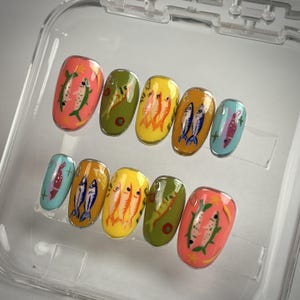 Könnte beinhalten: Ein Set aus zehn künstlichen Nägeln mit bunten Fischmotiven. Jeder Nagel hat ein anderes Design, darunter Fische in Orange-, Blau- und Grüntönen, vor Hintergründen in Rosa, Gelb und Türkis. Die Nägel sind oval geformt.