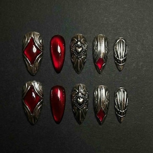 May include: A set of ten artificial nails with a gothic design. The nails feature a silver-colored metal base with intricate patterns and red jewel accents. Some nails have a heart design, while others have a diamond shape.