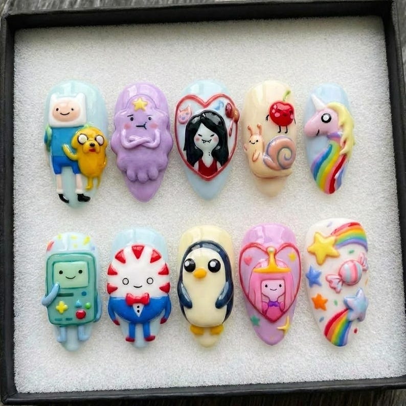 Puede incluir: Un conjunto de diez u&ntilde;as artificiales, cada una con dise&ntilde;os &uacute;nicos de personajes de dibujos animados y elementos coloridos. Los dise&ntilde;os incluyen un unicornio, un ping&uuml;ino y personajes de Adventure Time. Las u&ntilde;as est&aacute;n sobre un fondo blanco dentro de una caja negra.