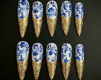 May include: Set of ten artificial nails with a white base and blue floral design. Each nail has a gold-colored, ornate tip. The nails are arranged on a black surface, showcasing a unique and artistic nail art design.