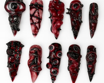 May include: A set of ten artificial nails, each with a distinctive design. The nails are long and pointed, with a dark red and black colour palette. They feature grotesque, organic, and eye-themed decorations. The nails are displayed on a white background.