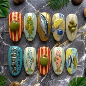Op de afbeelding: Een verzameling van tien uniek ontworpen nail art stukken. De ontwerpen omvatten vissen, olijven, citroenen en bosbessen. Eén nagel bevat de tekst "SARDINES À L'HUILE D'OLIVE". De nagels zijn geschilderd in verschillende kleuren, waaronder geel, rood, blauw en goud.
