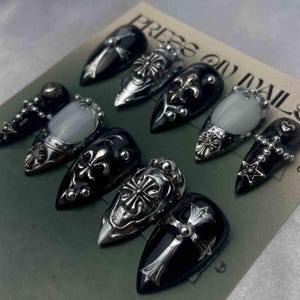 May include: A set of press-on nails with a gothic theme. The nails are black and feature silver-colored embellishments, including crosses, fleur-de-lis, and other ornate designs. Some nails have a pearlescent white stone.