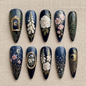 May include: A set of ten long, pointed, dark blue artificial nails. Each nail features intricate floral and celestial designs in white, gold, and pink. Designs include roses, foxgloves, moons, and stars, creating a detailed, artistic look.