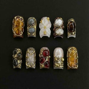 May include: A collection of ten ornate artificial nails. Each nail features a unique design with gold-colored metal accents and various embellishments, including gemstones, pearls, and carved floral elements. The nails are arranged on a dark surface.