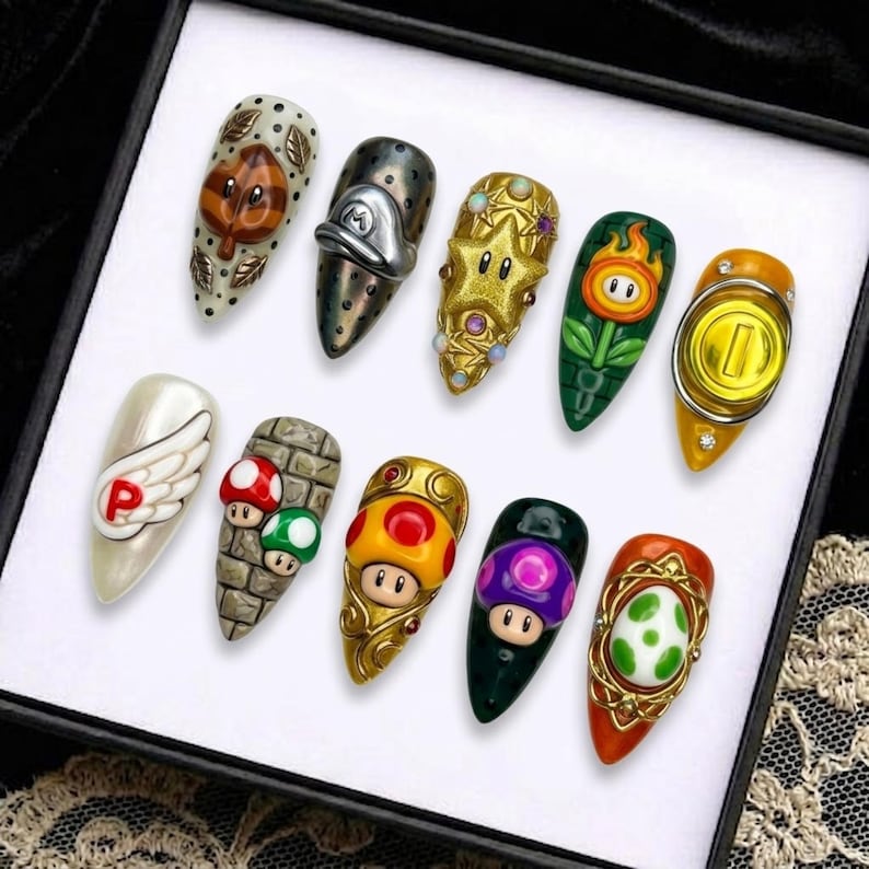 May include: A set of ten long, almond-shaped acrylic nails, each featuring a different design inspired by the Super Mario video game series. Designs include a leaf, a hat, a star, a fire flower, a coin, a wing, mushrooms, and more.