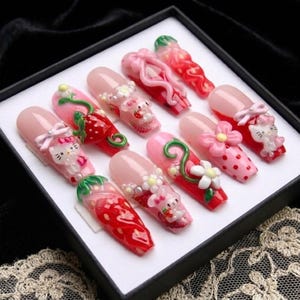 May include: A set of ten artificial nails, each with a unique design. The nails feature pink, red, and white colors, with decorative elements like strawberries, bows, flowers, and cartoon characters. The nails are presented in a black box.