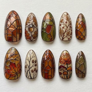 May include: A set of ten almond-shaped press-on nails featuring autumn-themed designs. Patterns include stained glass geometric grids in orange and green, detailed illustrations of acorns, oak leaves, maple leaves, berries, and mushrooms.
