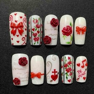 May include: A set of ten white artificial nails with various designs. Some nails feature red bows, roses, and hearts. Others have painted roses, musical notes, and the text "BE MINE".