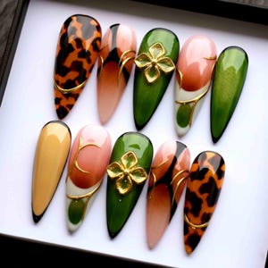 Può includere: Un set di dieci unghie finte lunghe a forma di mandorla. I disegni includono un motivo tartarugato, effetti sfumati, accenti floreali dorati e dettagli di linee dorate. I colori includono arancione, marrone, rosa, verde e giallo. Ogni unghia ha un bordo nero.