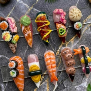 May include: A collection of ten long, pointed acrylic nails, each uniquely designed to resemble sushi and other Japanese food items. The nails feature detailed miniature sushi rolls, shrimp, and dumplings, with vibrant colors and textures.