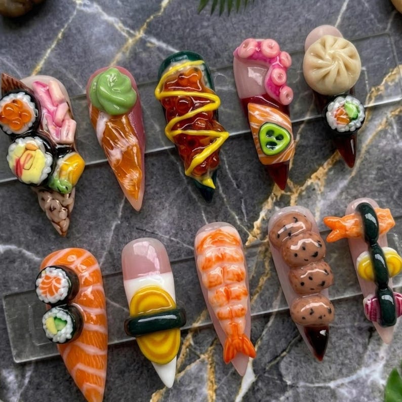 May include: A collection of ten long, pointed acrylic nails, each uniquely designed to resemble sushi and other Japanese food items. The nails feature detailed miniature sushi rolls, shrimp, and dumplings, with vibrant colors and textures.