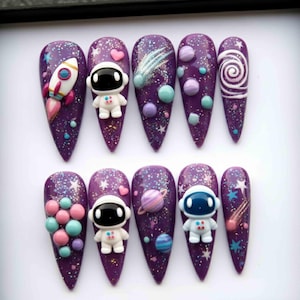 May include: A set of ten long, pointed, purple artificial nails decorated with space-themed designs. Each nail features glitter, stars, and various 3D elements like a rocket, astronaut, planets, and a comet. The nails are arranged in a neat row.