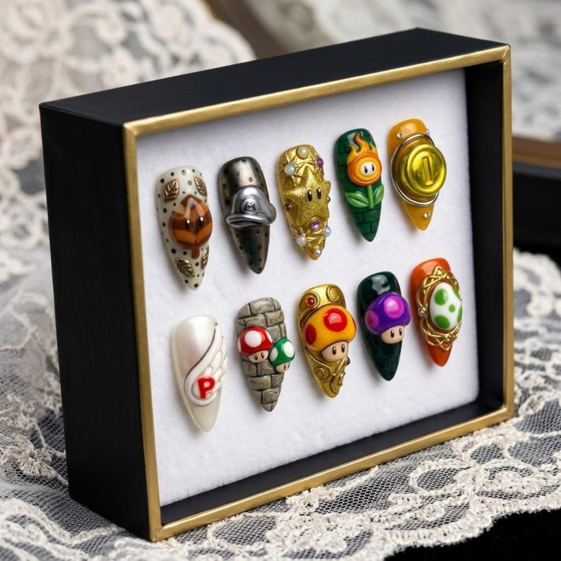 May include: A black framed display of ten custom-designed nail art pieces. Each nail features a different design inspired by the Super Mario video game series, including characters and iconic elements. The nails are in various colors and shapes.