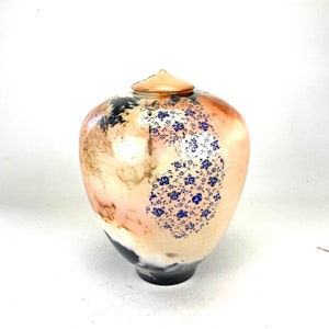 May include: A ceramic urn with a lid. The urn has a peach-coloured base with a dark blue floral pattern on one side. The top of the urn has a peach-coloured lid. The bottom of the urn is black.