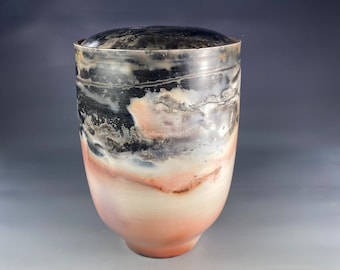 May include: A ceramic jar with a lid, featuring a black and white glaze with hints of pink and brown. The jar has a rounded body and a slightly flared lip.