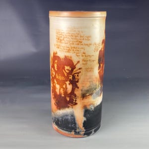 May include: A ceramic vase with a brown and white glaze. The vase has a Beatles image printed on it with handwritten lyrics around the image. The vase is approximately 10 inches tall.