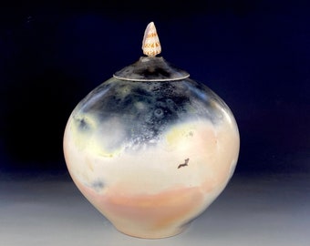 May include: A ceramic jar with a seashell lid. The jar is a light pink and white color with dark gray and black accents. The jar is round and has a smooth surface.