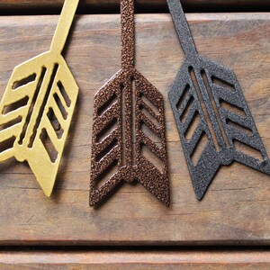 May include: Three metal arrowheads in different colors: gold, copper, and black. The arrowheads are arranged in a row on a wooden surface.