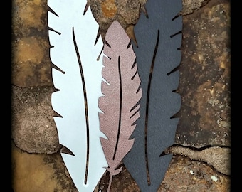 May include: Three metal feathers in different colors: white, brown, and black. The feathers are arranged vertically with the white feather on the left, the brown feather in the middle, and the black feather on the right. The feathers are all cut out of metal and have a smooth, polished finish.