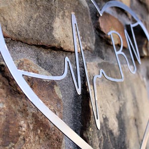May include: Metal heart-shaped wall decor with a stylised heartbeat and the letters "RN" inside. The decor is silver and is mounted on a stone wall.