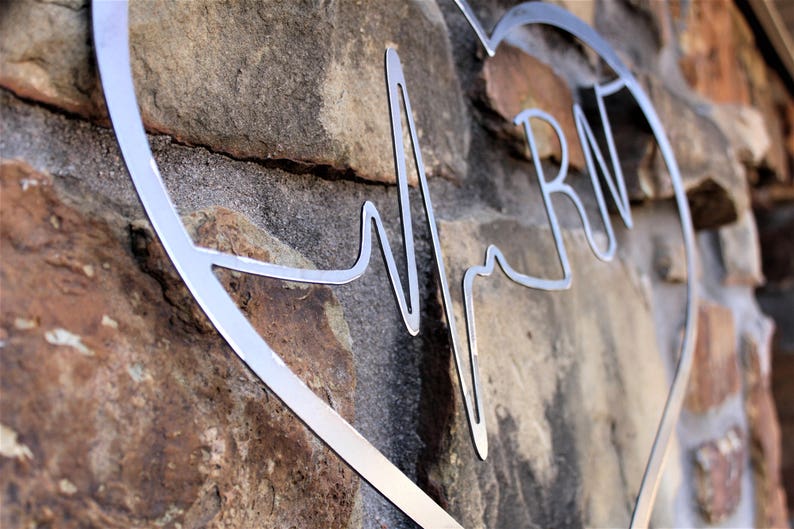 May include: Metal heart-shaped wall decor with a stylised heartbeat and the letters "RN" inside. The decor is silver and is mounted on a stone wall.