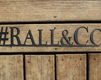 May include: A wooden sign with black metal letters spelling "#RALL&CO" on a wooden background.