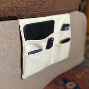 May include: A cream-colored fabric bedside storage organizer featuring multiple pockets hanging from the side of a bed frame. The pockets contain various items including a black tablet, a remote control, and small personal electronic devices.