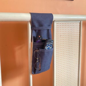 May include: A denim bed caddy with multiple pockets hangs from a white metal bed frame. The caddy holds two remote controls and a pair of eyeglasses. The background includes a poster and a decorative headboard.