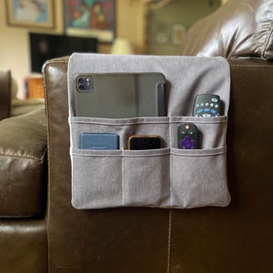 May include: A grey fabric armchair organizer featuring one large top pocket and three smaller bottom pockets. The organizer holds a tablet, a blue wallet, a smartphone, and two remote controls, draped over the side of a brown leather armchair.