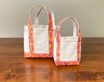 May include: Two canvas tote bags with white bodies and vibrant orange and pink patterned accents. The bags have matching handles and trim, and are displayed on a wooden surface.