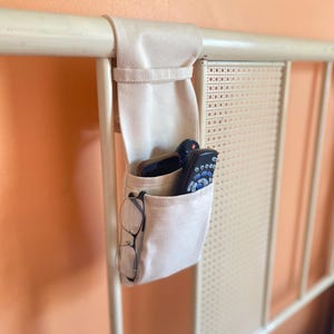 May include: A beige fabric bed organizer with multiple pockets hangs from a white metal headboard. The pockets hold a remote control, a smartphone, and a pair of eyeglasses. The headboard is against a peach-colored wall.