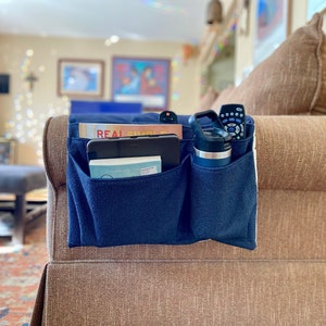 May include: A blue fabric organizer with multiple pockets hangs on the arm of a brown couch. The organizer holds a tablet, a book, a water bottle, and two remotes.