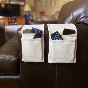 May include: Two off-white fabric armrest organizers draped over a brown leather sofa. Each organizer features two rectangular pockets containing black remote controls and smartphones, providing storage for small electronic devices and accessories.