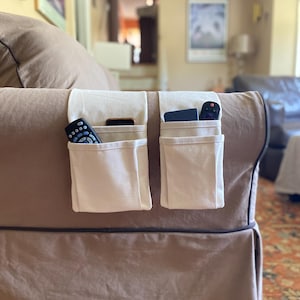 May include: Two off-white fabric armrest organizers draped over the side of a brown sofa. Each organizer features multiple vertical pockets containing various remote controls and a smartphone, providing storage for small electronic devices.