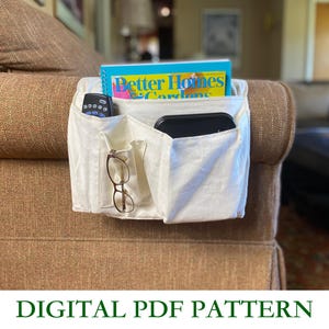 May include: A cream-colored couch arm organizer with multiple pockets, holding a remote, glasses, a black device, and a magazine titled "Better Homes & Gardens." The text "DIGITAL PDF PATTERN" is displayed below the organizer.