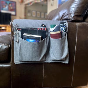 May include: A gray fabric organizer with multiple pockets attached to the arm of a brown leather armchair. The organizer holds a pair of glasses, a magazine, a book, a remote control, and a bottle of lotion.