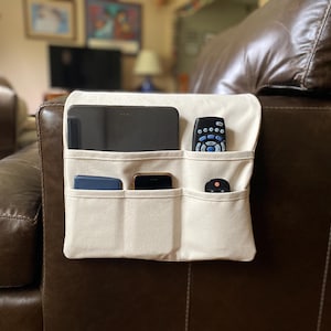 May include: A cream-colored fabric armchair organizer featuring five distinct pockets of varying sizes. The organizer is draped over the arm of a brown leather chair and holds a tablet, two remote controls, a smartphone, and a small blue rectangular object.