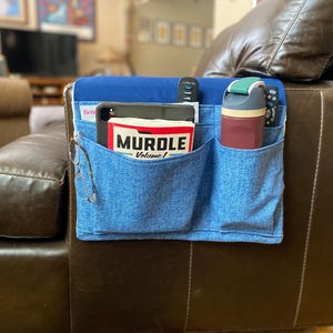 May include: A blue denim couch caddy with multiple pockets, holding a magazine, tablet, water bottle, remotes, and eyeglasses. The caddy is attached to a brown leather couch, providing convenient storage.