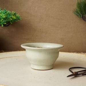 Może przedstawiać: Jasnozielona ceramiczna miska z szerokim brzegiem stoi na dużym, kremowym ceramicznym talerzu. Na talerzu spoczywa para małych, czarnych nożyczek. Dwa małe drzewka bonsai w brązowych donicach znajdują się w tle.