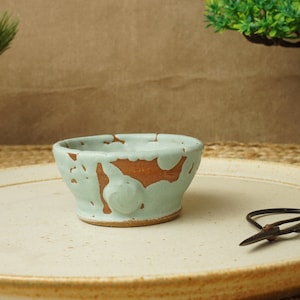 May include: A small, handmade ceramic bowl with a pale green glaze and exposed brown clay details. The bowl rests on a cream-coloured plate, with black scissors beside it. The bowl is likely for serving small portions of food.