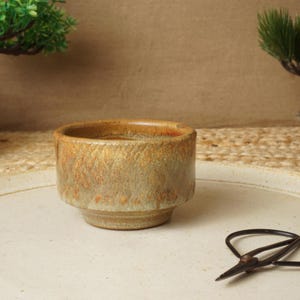 May include: A small, cylindrical ceramic bowl with a textured, mottled glaze in shades of earthy brown, tan, and muted green. The bowl features a slightly tapered base and a wide, open rim, showcasing a rustic, hand-crafted appearance.