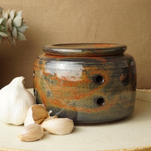 May include: A ceramic garlic keeper with a lid, glazed in brown and orange. The keeper has three ventilation holes. A whole garlic bulb and cloves are next to the keeper on a round plate. Decorative plants are in the background.