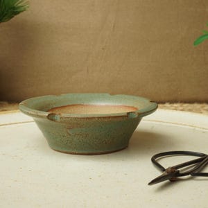 May include: A small, round, green ceramic bonsai pot with a scalloped rim. The interior is a warm, earthy tone. A pair of black scissors rests on the white surface in front of the pot. Decorative plants are in the background.