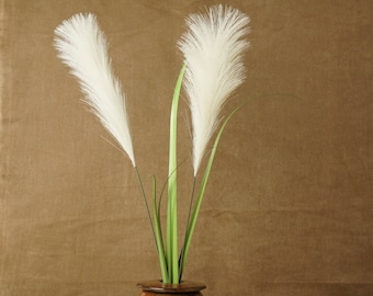 May include: Two white pampas grass plumes in a brown ceramic vase with green stems. The vase is sitting on a woven straw mat.